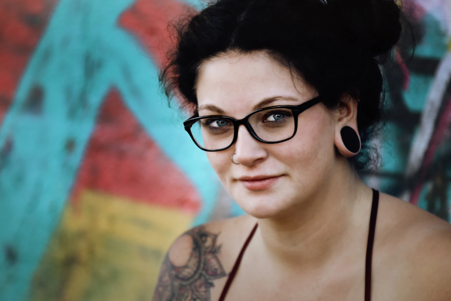 A tattooed woman in heavy cat-eye frames standing in front of a painted wall