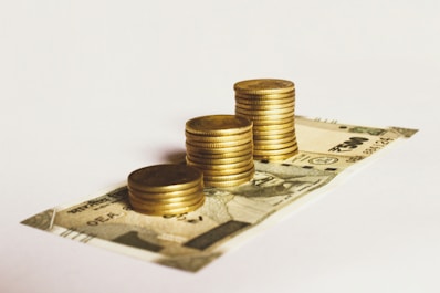 A stack of gold coins is neatly placed on top of a 500 Indian rupee banknote. The arrangement suggests a focus on wealth accumulation or savings. The background is plain and minimalistic, drawing attention to the currency and coins.