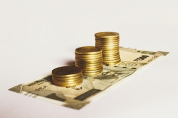 A stack of gold coins is neatly placed on top of a 500 Indian rupee banknote. The arrangement suggests a focus on wealth accumulation or savings. The background is plain and minimalistic, drawing attention to the currency and coins.