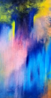 A vibrant abstract painting featuring a blend of colors including deep blue, purple, pink, yellow, and hints of green. The colors appear to merge and bleed into each other, creating a fluid, dreamlike effect. The top section of the image is darker, with intense cloud-like formations, while the bottom half transitions into lighter, more pastel shades.