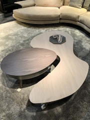 A modern living room setup with a curved beige sofa positioned on a textured grey carpet. In front of the sofa, there are two asymmetrical coffee tables, one light wooden and the other dark, holding a few glasses on a tray.