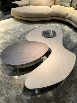 A modern living room setup with a curved beige sofa positioned on a textured grey carpet. In front of the sofa, there are two asymmetrical coffee tables, one light wooden and the other dark, holding a few glasses on a tray.