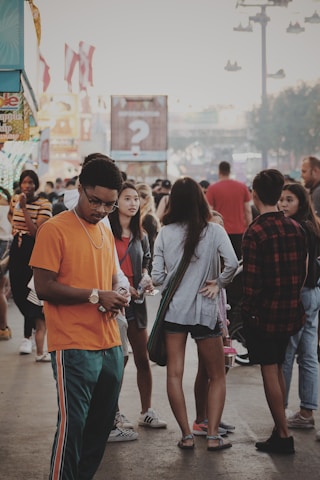 A busy outdoor scene with a diverse group of people at what appears to be a fair or public event. The background includes stands with signage and colorful decorations. A young man in an orange shirt is looking at his phone in the foreground, while behind him, other individuals are engaged in conversation or walking.