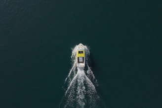 Aerial view of a boat with a streamlined design moving through dark water, leaving a symmetric wake behind. The boat has a predominantly white structure with a yellow top section.