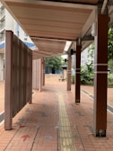 A covered walkway with a tiled floor and wooden beams overhead extends forward. The path includes a tactile guiding strip for accessibility. Greenery, such as bushes and trees, lines the side, and urban buildings are visible in the background.