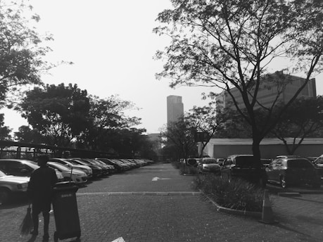 A black and white photo of a parking lot lined with cars and bordered by trees. In the foreground, a person walks with a push cart and a broom. Skyscrapers are visible in the background beyond the trees.