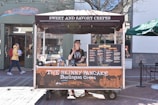 A crepe cart named 'The Skinny Pancake' is stationed outdoors with a person standing in it. The cart offers sweet and savory crepes and features a menu board displaying various options. A passerby in a yellow jacket and red backpack is visible on the left, walking past the storefront of a shop called Zinnia. In the background, there is a Starbucks sign and other shops on a brick pedestrian area.