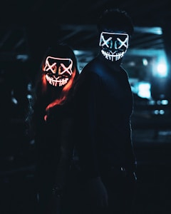 Two individuals stand in a dimly lit environment, wearing glowing LED masks with stitched mouth designs and X-shaped eyes. The masks emit bright neon colors, one in red and the other in blue, creating an eerie and mysterious atmosphere.