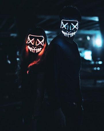 Two individuals stand in a dimly lit environment, wearing glowing LED masks with stitched mouth designs and X-shaped eyes. The masks emit bright neon colors, one in red and the other in blue, creating an eerie and mysterious atmosphere.