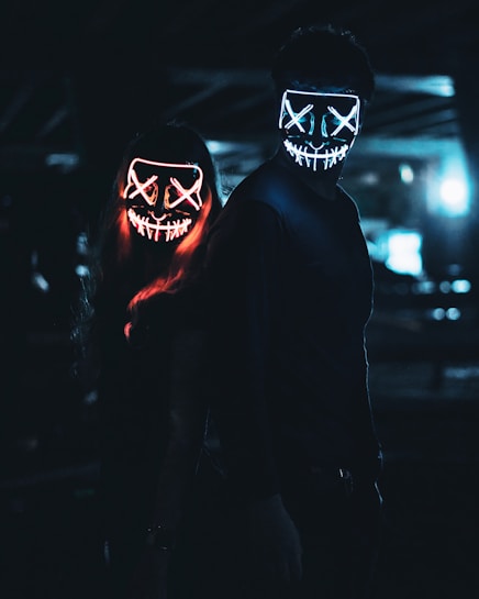 Two individuals stand in a dimly lit environment, wearing glowing LED masks with stitched mouth designs and X-shaped eyes. The masks emit bright neon colors, one in red and the other in blue, creating an eerie and mysterious atmosphere.