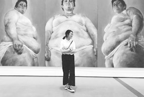 A person stands in front of a large triptych artwork depicting three larger-than-life, full-body portraits in grayscale. The paintings feature three figures, each in a similar pose, wearing minimal clothing. The individual observing the artwork is dressed casually and appears contemplative.