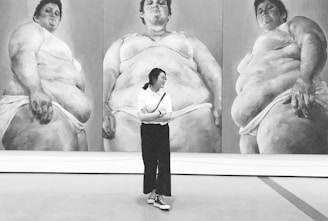 A person stands in front of a large triptych artwork depicting three larger-than-life, full-body portraits in grayscale. The paintings feature three figures, each in a similar pose, wearing minimal clothing. The individual observing the artwork is dressed casually and appears contemplative.