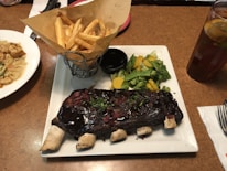 A plate with a rack of ribs covered in a dark sauce, garnished with herbs and minced onions. To the side are cooked vegetables including broccoli, yellow squash, and snow peas. A basket of seasoned fries sits beside the ribs, and there is a small cup of sauce or dip. In the background, a glass of iced tea with lemon is present, along with another dish containing pasta and grilled shrimp.