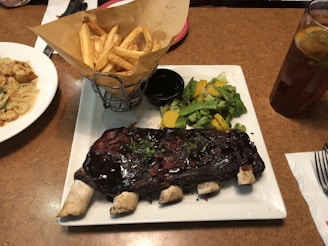 A plate with a rack of ribs covered in a dark sauce, garnished with herbs and minced onions. To the side are cooked vegetables including broccoli, yellow squash, and snow peas. A basket of seasoned fries sits beside the ribs, and there is a small cup of sauce or dip. In the background, a glass of iced tea with lemon is present, along with another dish containing pasta and grilled shrimp.