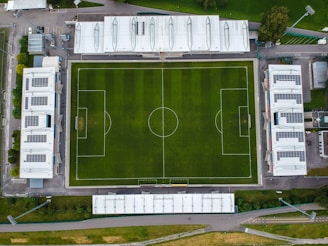 An aerial view of a rectangular soccer field with clearly marked lines, two goals, and surrounded by several buildings. The grass appears lush and well-maintained, and the surrounding structures include large white-roofed buildings.