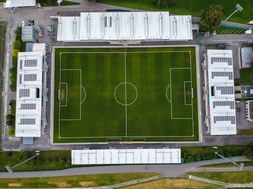 An aerial view of a rectangular soccer field with clearly marked lines, two goals, and surrounded by several buildings. The grass appears lush and well-maintained, and the surrounding structures include large white-roofed buildings.