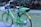 A green electric bike with a basket and labeled 'Lime-E' is parked on a paved path in a grassy park. The bike has a prominent yellow rear wheel cover and is equipped with a motor on the rear wheel.