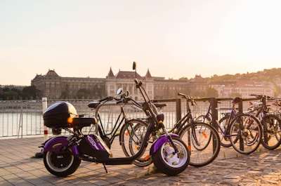 Multiple bicycles and an electric scooter are parked on a cobblestone path by a railing overlooking a large river. In the background, a grand historic building is visible across the river under a clear, pale sky as the sun sets, casting a warm glow over the scene.