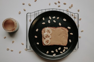 A piece of toast topped with creamy peanut butter and sliced bananas rests on a black plate. The plate is placed on a cooling rack with scattered peanuts around it. A jar of peanut butter is visible to the left, partly open. The background is a plain white surface.