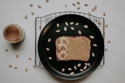 A piece of toast topped with creamy peanut butter and sliced bananas rests on a black plate. The plate is placed on a cooling rack with scattered peanuts around it. A jar of peanut butter is visible to the left, partly open. The background is a plain white surface.
