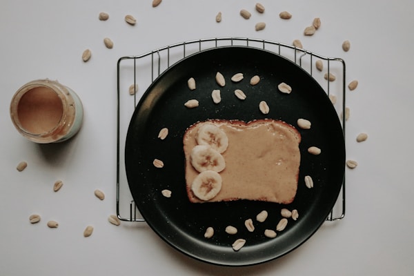 A piece of toast topped with creamy peanut butter and sliced bananas rests on a black plate. The plate is placed on a cooling rack with scattered peanuts around it. A jar of peanut butter is visible to the left, partly open. The background is a plain white surface.
