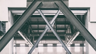 A structural steel framework in an industrial setting, characterized by large, overlapping beams forming an X pattern. The setting appears to be inside a warehouse or factory with visible metal rafters and ceiling in the background.