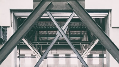 A structural steel framework in an industrial setting, characterized by large, overlapping beams forming an X pattern. The setting appears to be inside a warehouse or factory with visible metal rafters and ceiling in the background.