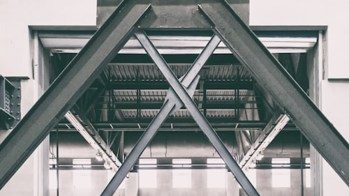 A structural steel framework in an industrial setting, characterized by large, overlapping beams forming an X pattern. The setting appears to be inside a warehouse or factory with visible metal rafters and ceiling in the background.