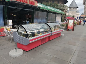 An outdoor food display featuring two large, red, refrigerated display cases filled with various dishes and beverages. These display cases are situated on a tiled walkway in front of a cafe or restaurant. The cafe has a canopy with green awnings and is part of a larger architectural environment with castle-like buildings in the background. There are some people walking in the distance and a few tables and chairs to the side of the display cases.