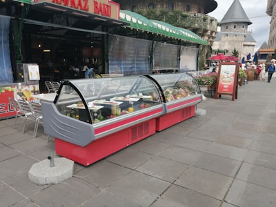 An outdoor food display featuring two large, red, refrigerated display cases filled with various dishes and beverages. These display cases are situated on a tiled walkway in front of a cafe or restaurant. The cafe has a canopy with green awnings and is part of a larger architectural environment with castle-like buildings in the background. There are some people walking in the distance and a few tables and chairs to the side of the display cases.