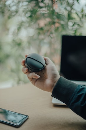 A hand holds a round, black electronic device with a logo. A blurred background of green leaves creates an outdoor atmosphere. A smartphone lies on a wooden table, and an open laptop is partly visible.