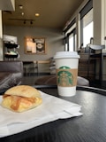 A cozy cafe interior with a focus on a round black table holding a paper-wrapped breakfast sandwich and a white takeaway coffee cup with a brown sleeve displaying the Starbucks logo. In the background, brown leather chairs and tables are arranged, with a large window letting in natural light.