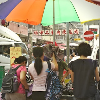 A busy street market scene with several people browsing. Overhead, a large multicolored umbrella provides shade. In the background, signs with Chinese characters are visible, and a white van is parked nearby.