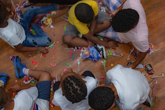 Children are sitting on a wooden floor surrounded by scattered candy wrappers and candy. They are reaching out to grab and sort the candies.