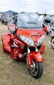 A bright red Honda trike motorcycle is parked on grass, surrounded by classic cars at what seems to be an outdoor vehicle show. The motorcycle has a large windscreen and chrome details. People can be seen in the background, indicating a gathering or event.