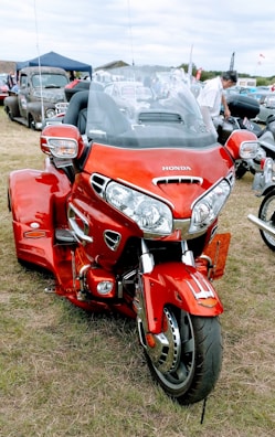 A bright red Honda trike motorcycle is parked on grass, surrounded by classic cars at what seems to be an outdoor vehicle show. The motorcycle has a large windscreen and chrome details. People can be seen in the background, indicating a gathering or event.