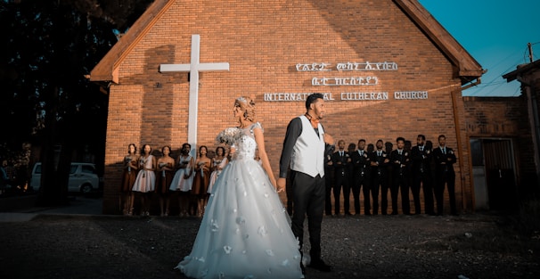 A wedding couple stands in front of a brick church with a large white cross. The bride wears a white dress adorned with flowers and the groom is in a black suit with a white vest. Behind them, bridesmaids and groomsmen are lined up against the church wall.