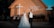 A wedding couple stands in front of a brick church with a large white cross. The bride wears a white dress adorned with flowers and the groom is in a black suit with a white vest. Behind them, bridesmaids and groomsmen are lined up against the church wall.