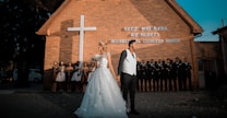 A wedding couple stands in front of a brick church with a large white cross. The bride wears a white dress adorned with flowers and the groom is in a black suit with a white vest. Behind them, bridesmaids and groomsmen are lined up against the church wall.