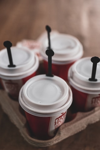 Four paper coffee cups with white lids and red sleeves are placed in a cardboard cup holder. Each cup has a black stirrer inserted into the lid.