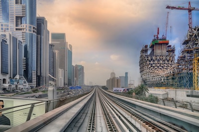 A modern cityscape with futuristic skyscrapers lining the left side and an intricate building under construction on the right. A railway track runs straight through the city, leading towards the distant skyline. The sky is partly cloudy with a warm hue.