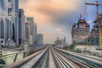 A modern cityscape with futuristic skyscrapers lining the left side and an intricate building under construction on the right. A railway track runs straight through the city, leading towards the distant skyline. The sky is partly cloudy with a warm hue.