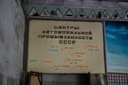 A wall displays a vintage sign written in Russian, featuring the words related to automotive industry centers in the USSR. The sign includes the names of various cities and automobile manufacturers. Above the sign, there are red-bordered triangular warning signs with various symbols.
