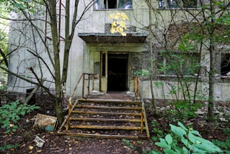 An abandoned, derelict building surrounded by overgrown vegetation. The front door is open, leading to a dark interior, and the exterior is worn with noticeable decay. Rusty railings and steps lead up to the entrance, while trees and bushes obscure parts of the scene.