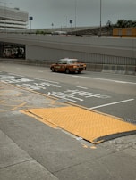 A taxi travels on a multi-lane road with Asian characters painted on the pavement, suggesting traffic instructions. The road is lined with barriers and is situated under an overpass. In the background, there is a multi-story parking structure, and various signs are visible. The sky appears overcast, and there is a pedestrian sidewalk beside the road with a textured yellow ramp.