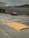 A taxi travels on a multi-lane road with Asian characters painted on the pavement, suggesting traffic instructions. The road is lined with barriers and is situated under an overpass. In the background, there is a multi-story parking structure, and various signs are visible. The sky appears overcast, and there is a pedestrian sidewalk beside the road with a textured yellow ramp.
