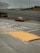 A taxi travels on a multi-lane road with Asian characters painted on the pavement, suggesting traffic instructions. The road is lined with barriers and is situated under an overpass. In the background, there is a multi-story parking structure, and various signs are visible. The sky appears overcast, and there is a pedestrian sidewalk beside the road with a textured yellow ramp.