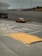 A taxi travels on a multi-lane road with Asian characters painted on the pavement, suggesting traffic instructions. The road is lined with barriers and is situated under an overpass. In the background, there is a multi-story parking structure, and various signs are visible. The sky appears overcast, and there is a pedestrian sidewalk beside the road with a textured yellow ramp.