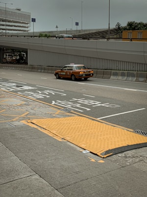 A taxi travels on a multi-lane road with Asian characters painted on the pavement, suggesting traffic instructions. The road is lined with barriers and is situated under an overpass. In the background, there is a multi-story parking structure, and various signs are visible. The sky appears overcast, and there is a pedestrian sidewalk beside the road with a textured yellow ramp.