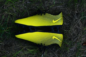 Two bright yellow and black soccer cleats with a textured pattern, placed against a backdrop of grass.
